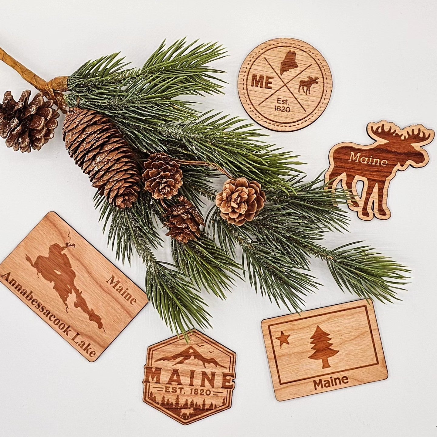 Beebow's laser-cut wooden magnets featuring Maine illustrations scattered around a large pine bough with pinecones