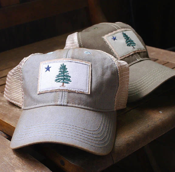 Slightly distressed baseball hats in neutral colors featuring the 1901 Maine Flag, in natural lighting on a rustic wooden chair
