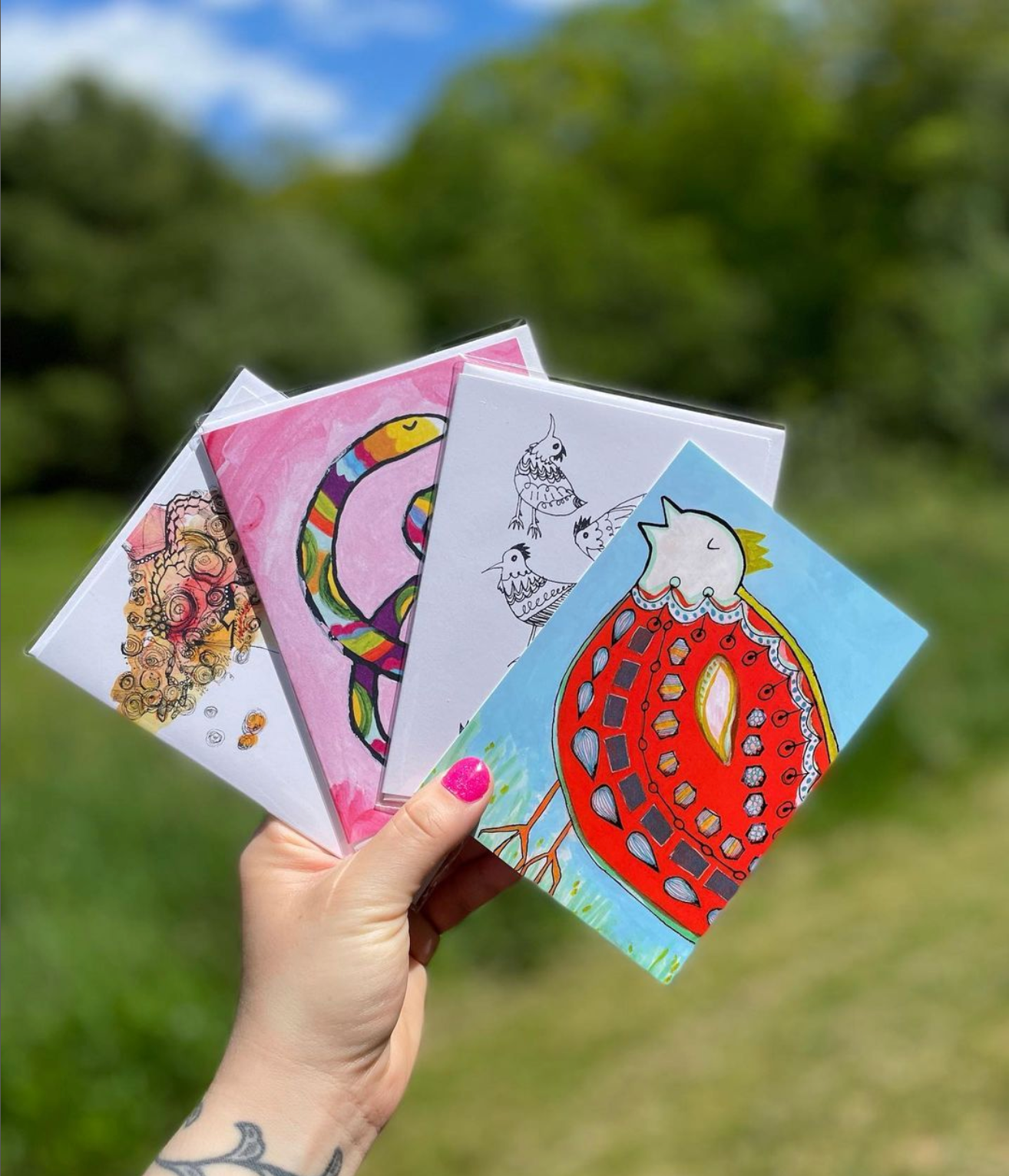 Hana Firestone colorful illustrated greeting cards held by a hand with pink nails and tattoos against a background of green trees and a blue sky