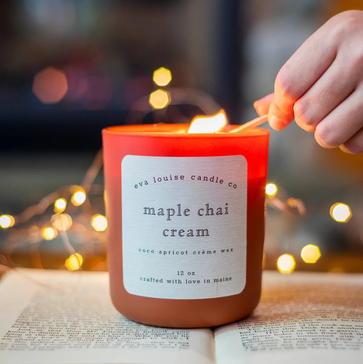 Eva Louie Candles' Maple Chai Cream Candle being lit with a match, while resting on an open book with cozy glowing fairy lights in the background